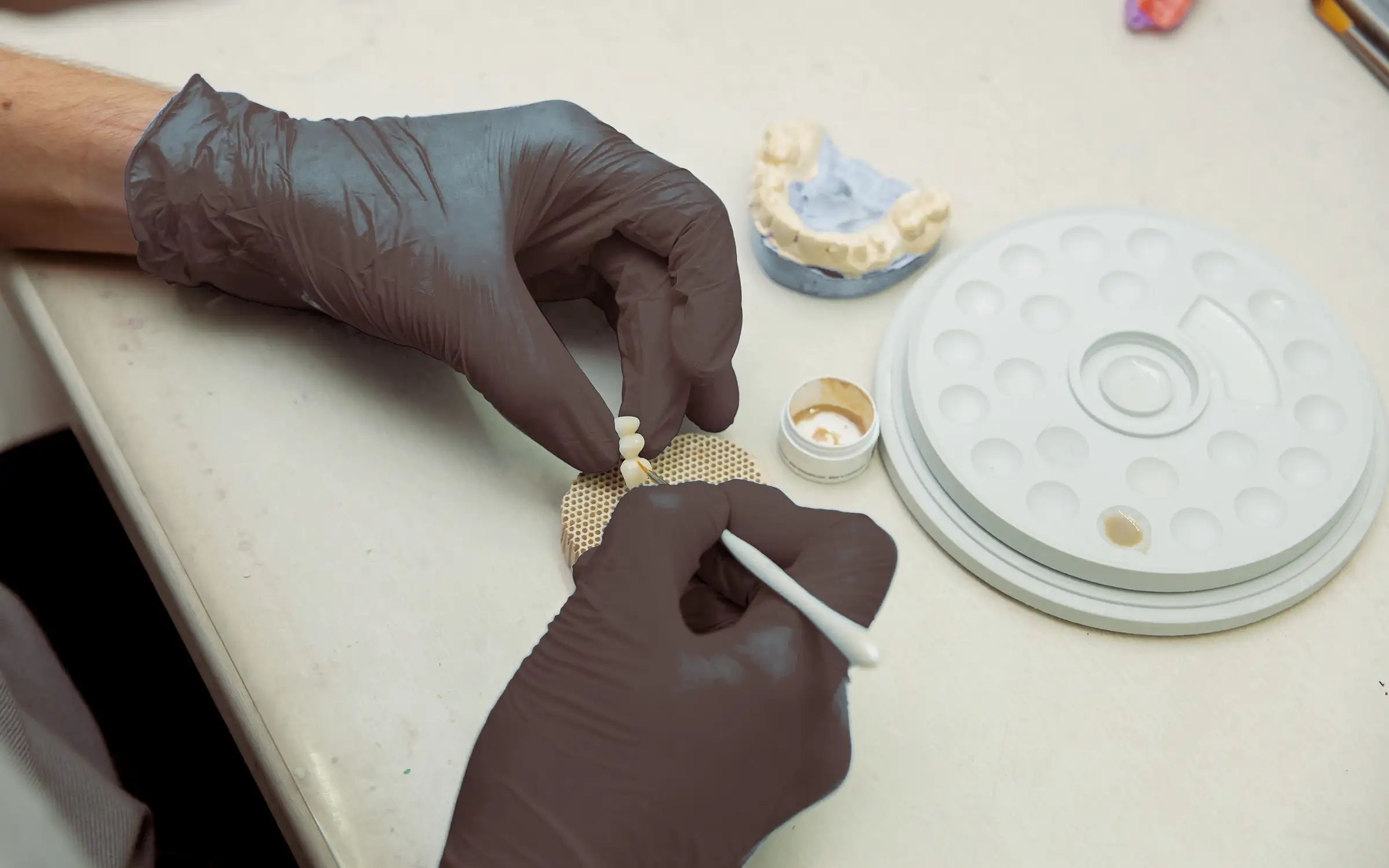 Mãos de um técnico em prótese aplicando cerâmica em uma pequena ponte de dentes, com um modelo de gesso ao fundo